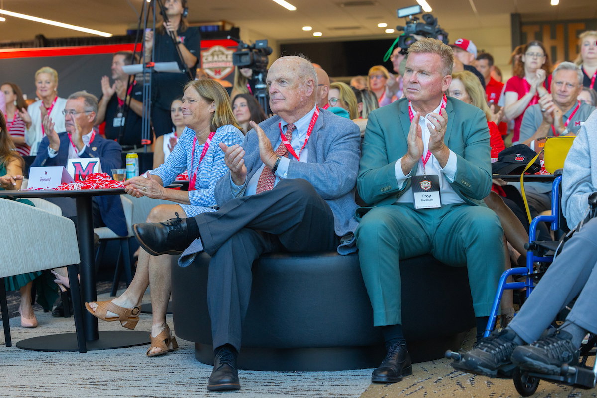 Several attendees, including one identified as Troy, are seated and applauding at an event with cameras and a branded backdrop.