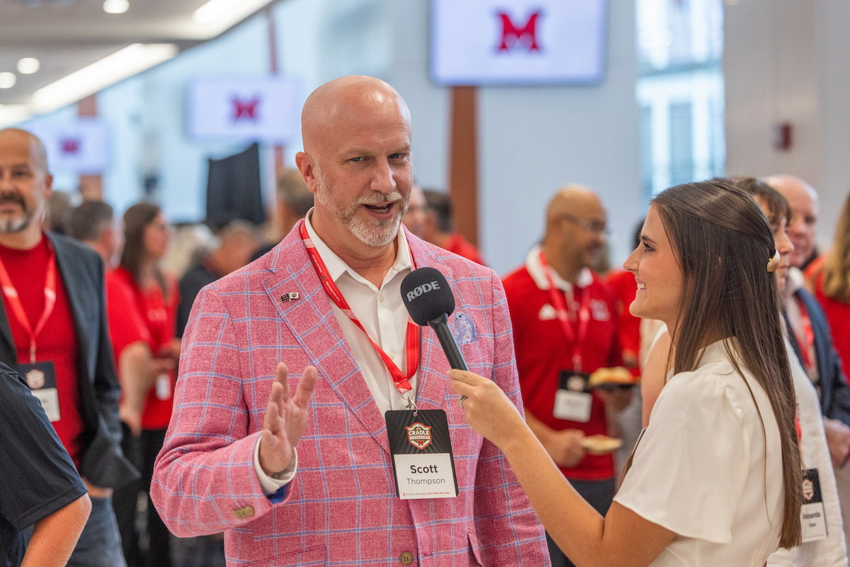 A person in a pink plaid jacket is being interviewed by a reporter holding a Rode microphone at an event with multiple attendees in the background.