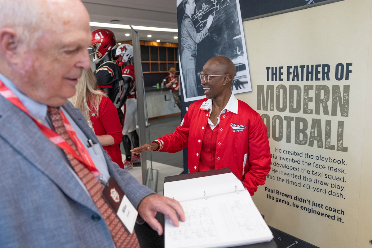 A person in a red jacket gestures toward a display honoring Paul Brown, the “Father of Modern Football,” while another individual reviews documents nearby.