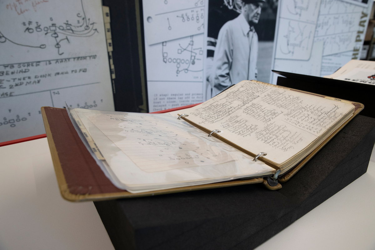 An open, aged binder filled with handwritten notes and diagrams sits on a display stand, surrounded by football strategy charts and a black-and-white photograph.